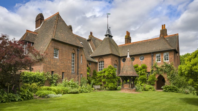 The imposing red facade of the house at Red House in London with its well and high peaked roof surrounded by a luscious green garden full of plants and flowers